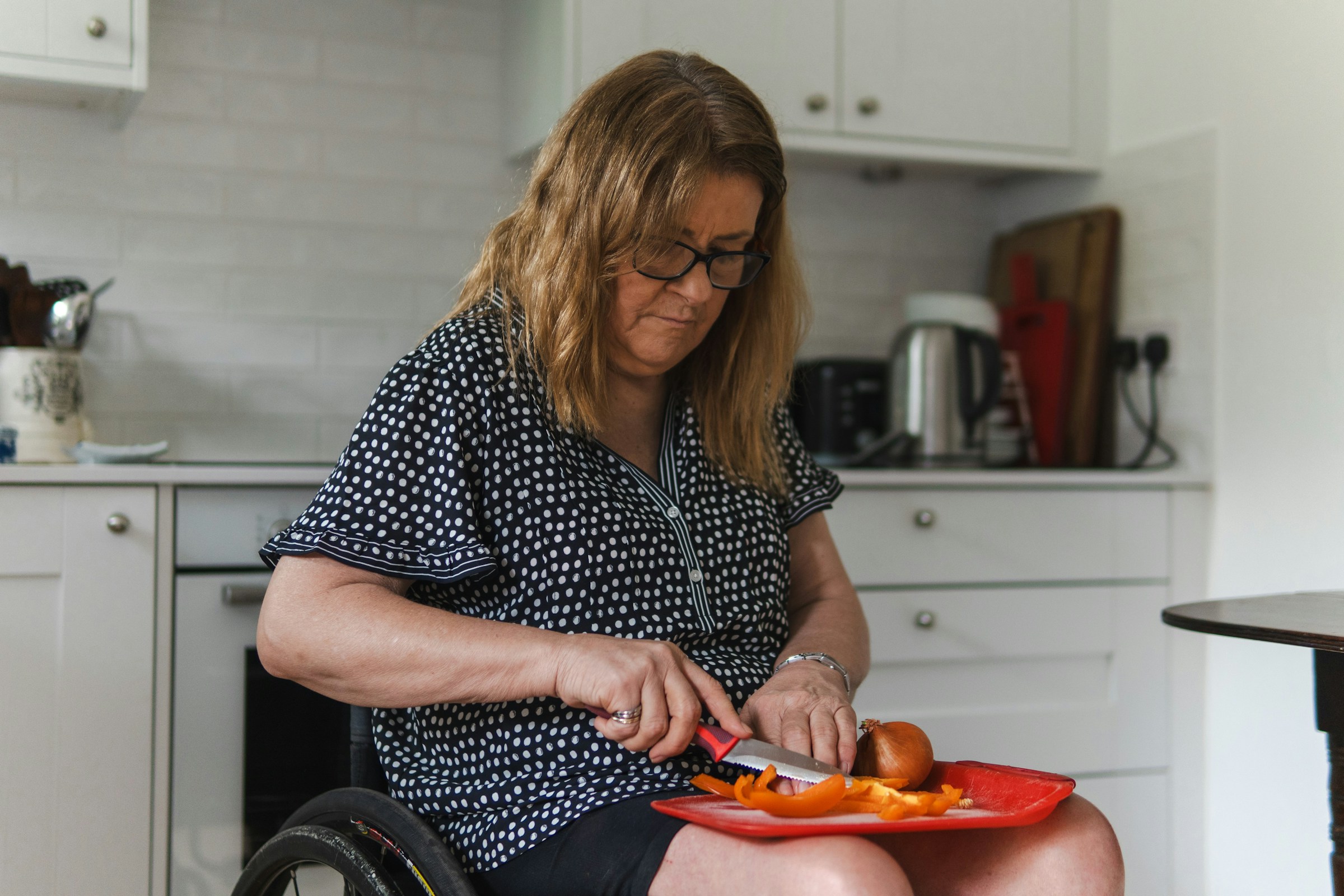 A woman in a wheelchair slicing vegetables on a lap tray in her own kitchen