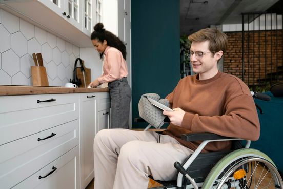 A wheelchair user reading a tablet at his kitchen table while another person prepares food at the counter behind him