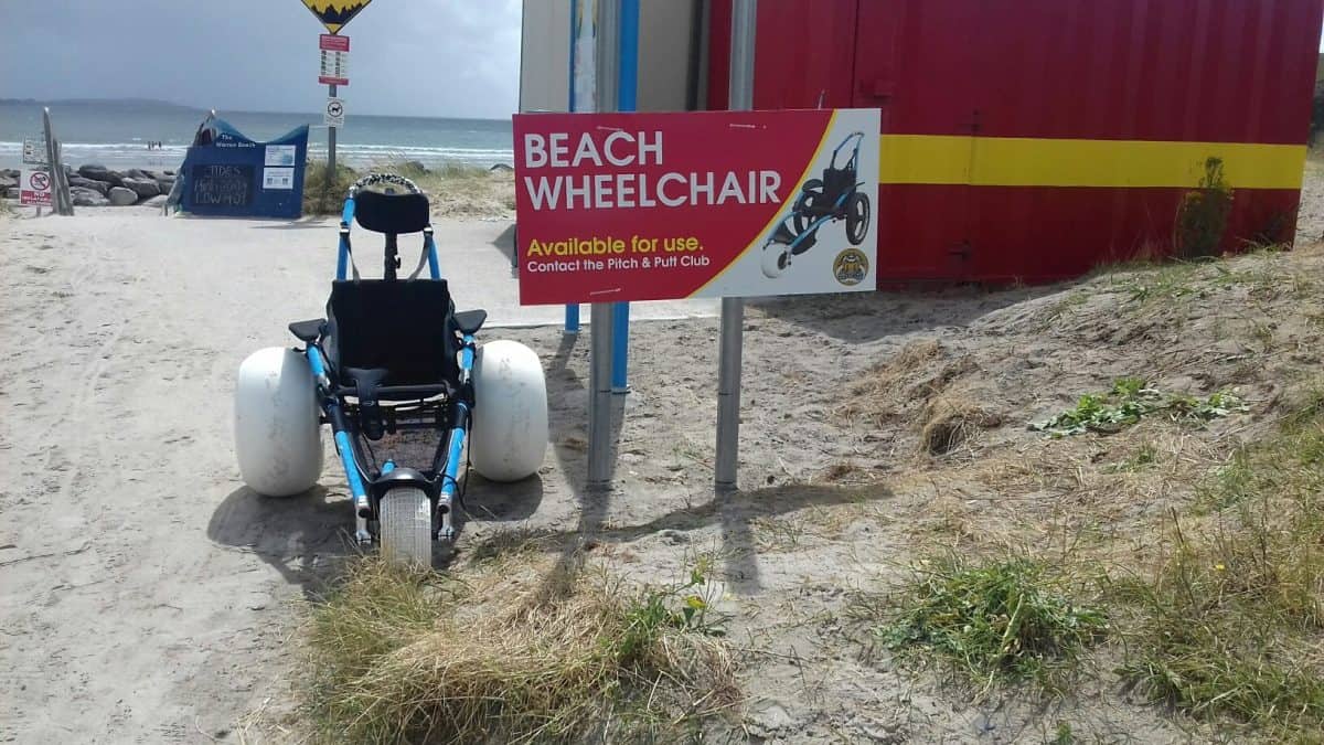 Photo of a wheelchair on the beach next to a sign that says Beach Wheelchair. Available for Use. Contact the Pitch & Putt Club